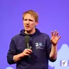 Nicolas Grekas with a mic in his right hand raising his left hand on stage at SymfonyCon Amsterdam 2025